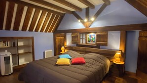 Un dormitorio con techo inclinado, vigas de madera, una cama con muchos almohadones, lámparas de noche y un radiador.