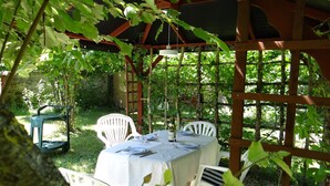 Un área de comedor al aire libre con mesa y sillas cubiertas, rodeada de follaje y una pérgola de madera.