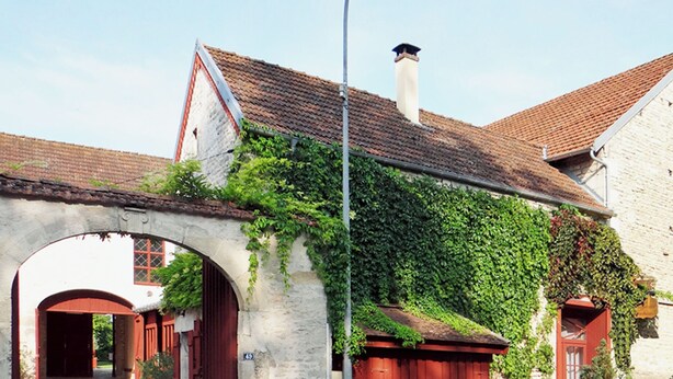 Un edificio de piedra con puerta roja y un porche cubierto, rodeado de vegetación y un cielo despejado.