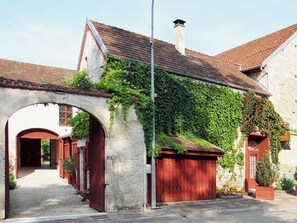 Ein Steinhaus mit roter Tür und einer überdachten Eingangspforte, umgeben von viel Grün und blauem Himmel.