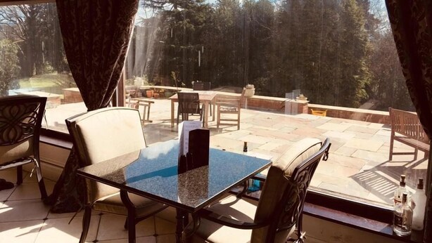 A sunlit patio with a glass table and chairs, overlooking a garden and a forested area. - Brass Castle Country House Accommodation (Middlesbrough)