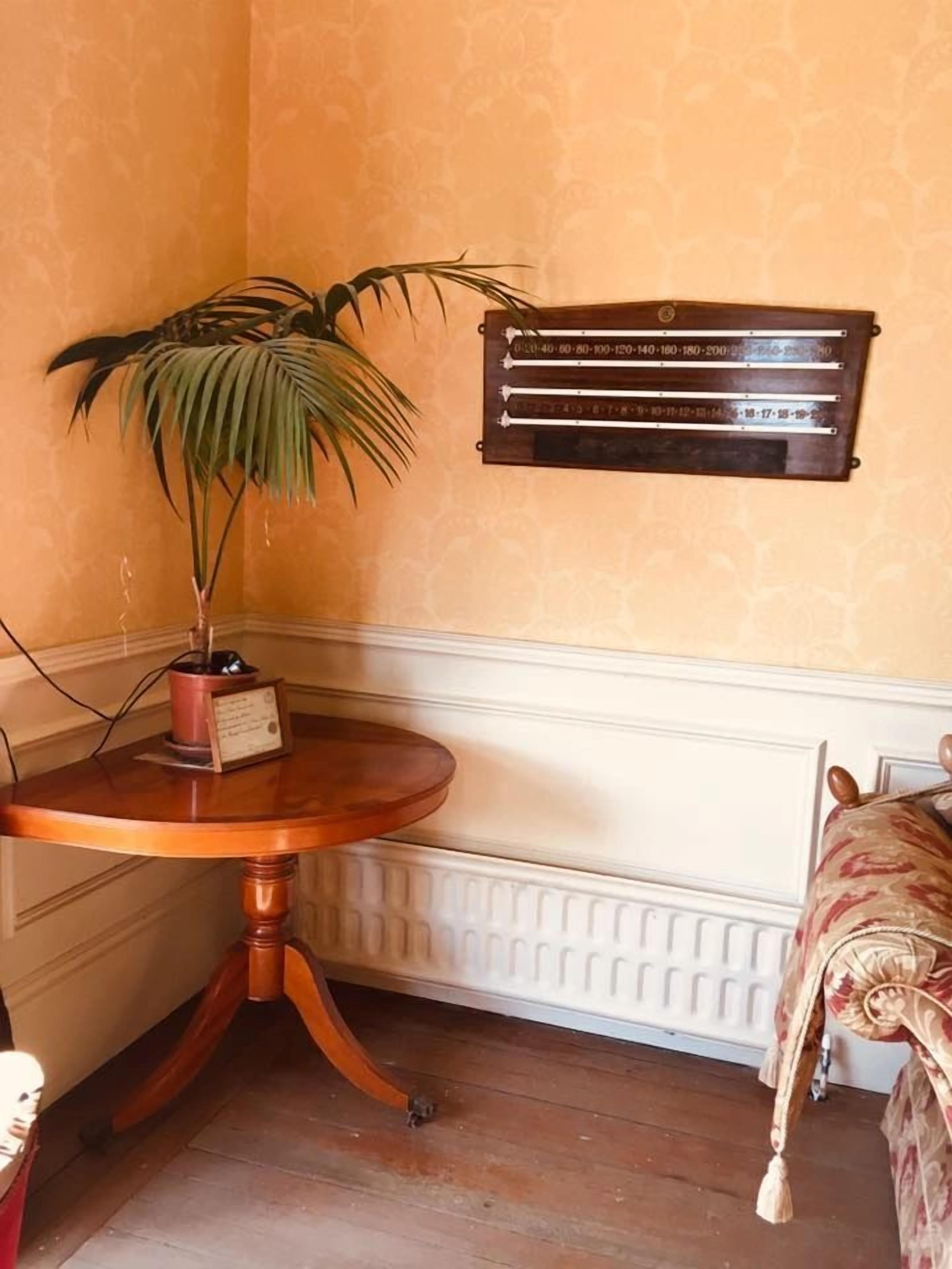 A corner room with a wooden table, a potted plant, a radiator, and a bed with a patterned bedspread.
