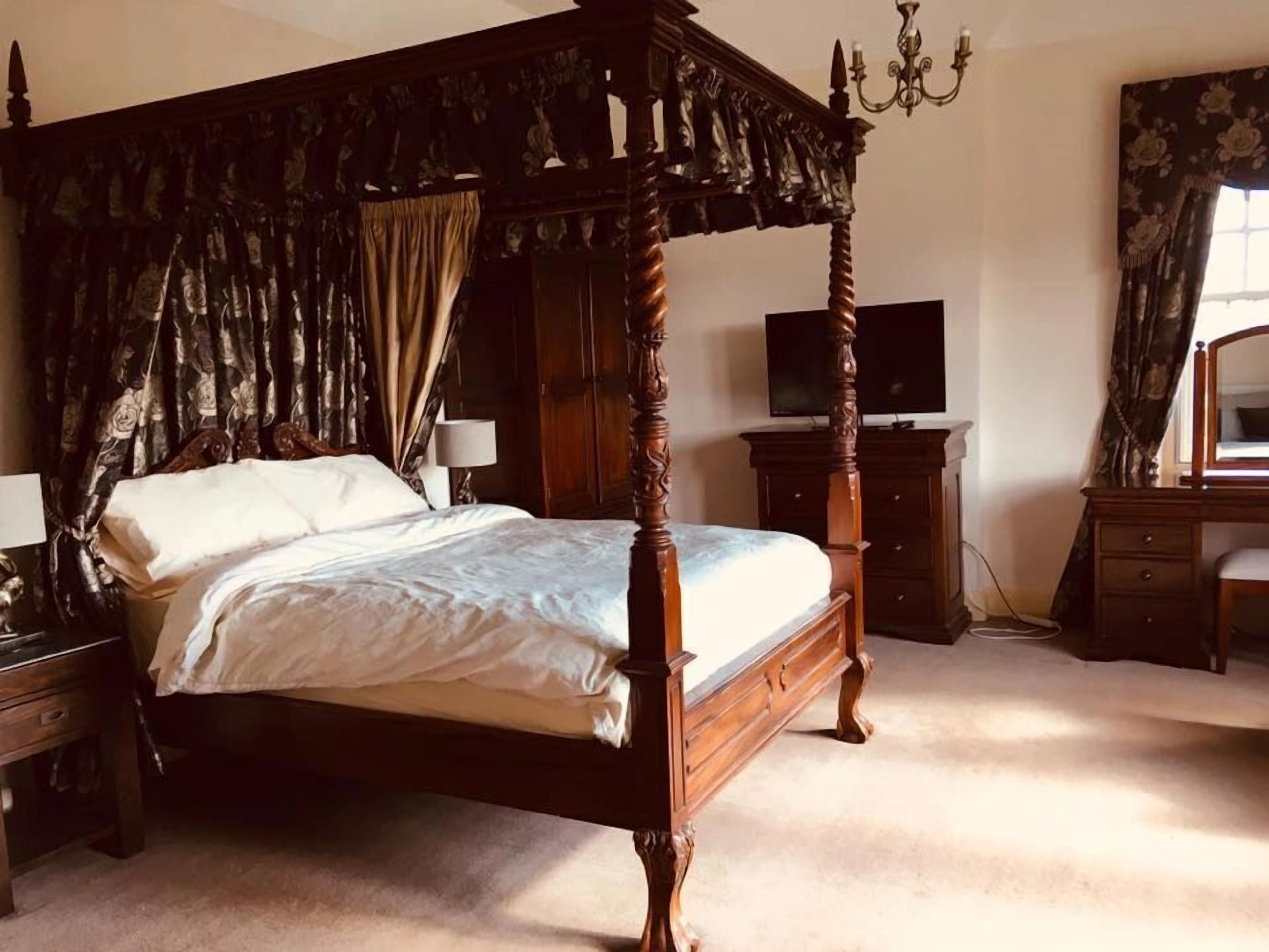 A four-poster canopy bed with a television on a dresser, a desk, and a chair in a bedroom.
