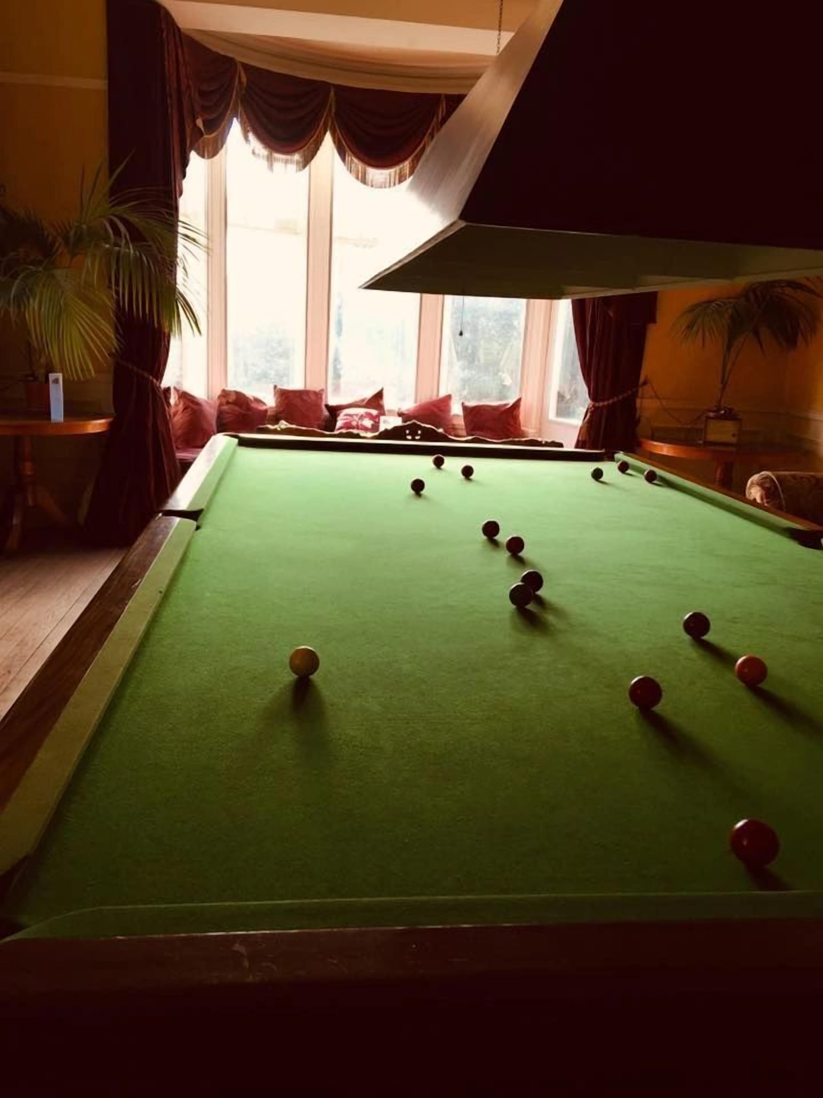 A pool table with balls in a room with large windows, a sofa with red cushions, and potted plants.
