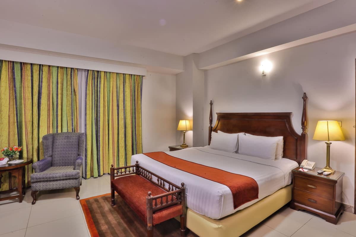 Une chambre d’hôtel avec un grand lit, une chaise, une table de chevet avec une lampe et une fenêtre avec des rideaux.