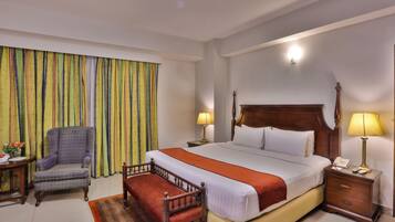 A hotel room with a large bed, a chair, a bedside table with a lamp, and a window with curtains.