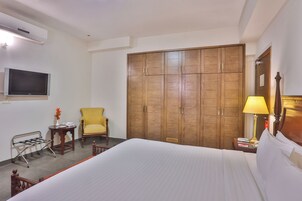 Quarto de hotel com cama, guarda-roupa de madeira, televisão, cadeira, mesa de cabeceira com abajur e uma pequena mesa com um vaso.