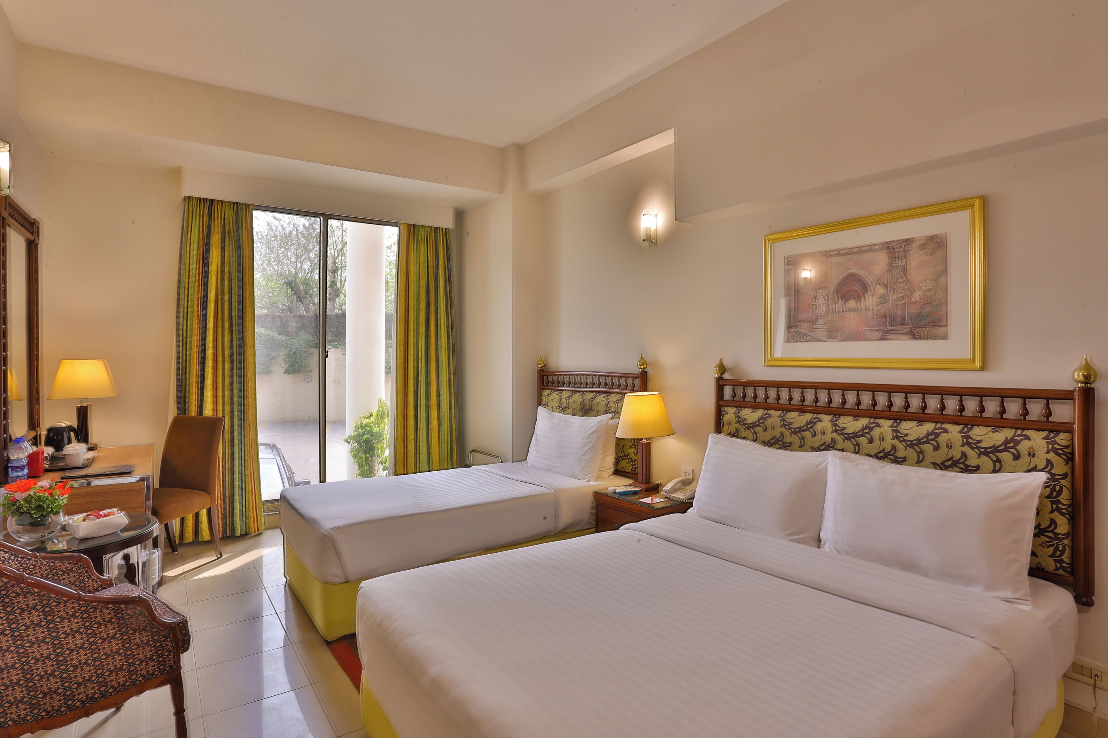 Habitación de hotel con dos camas, un escritorio con lámpara, una silla, una ventana con cortinas y un cuadro en la pared.