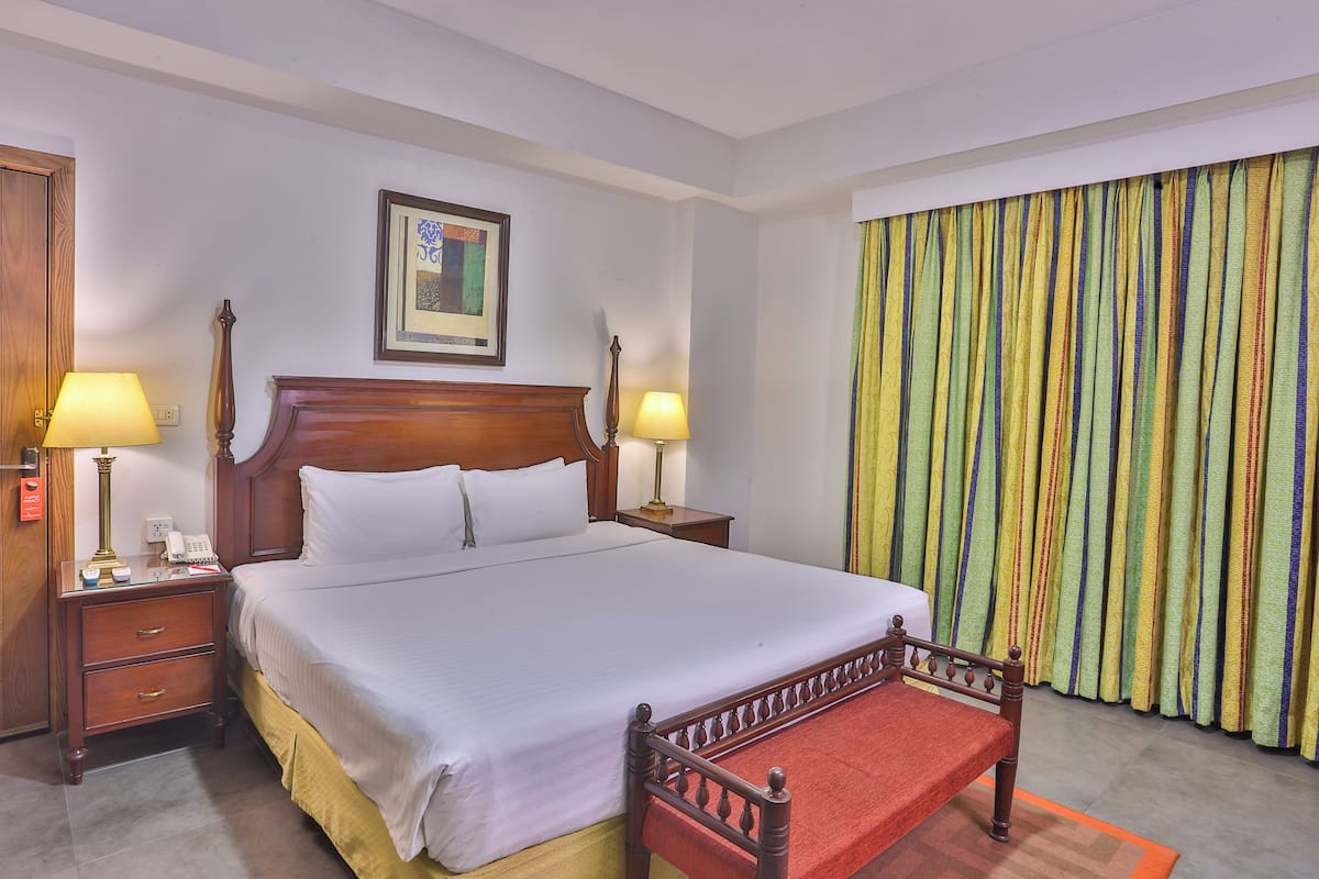 Une chambre d’hôtel avec un grand lit, deux tables de chevet avec des lampes, une tête de lit en bois, un tableau encadré au mur et un rideau coloré.