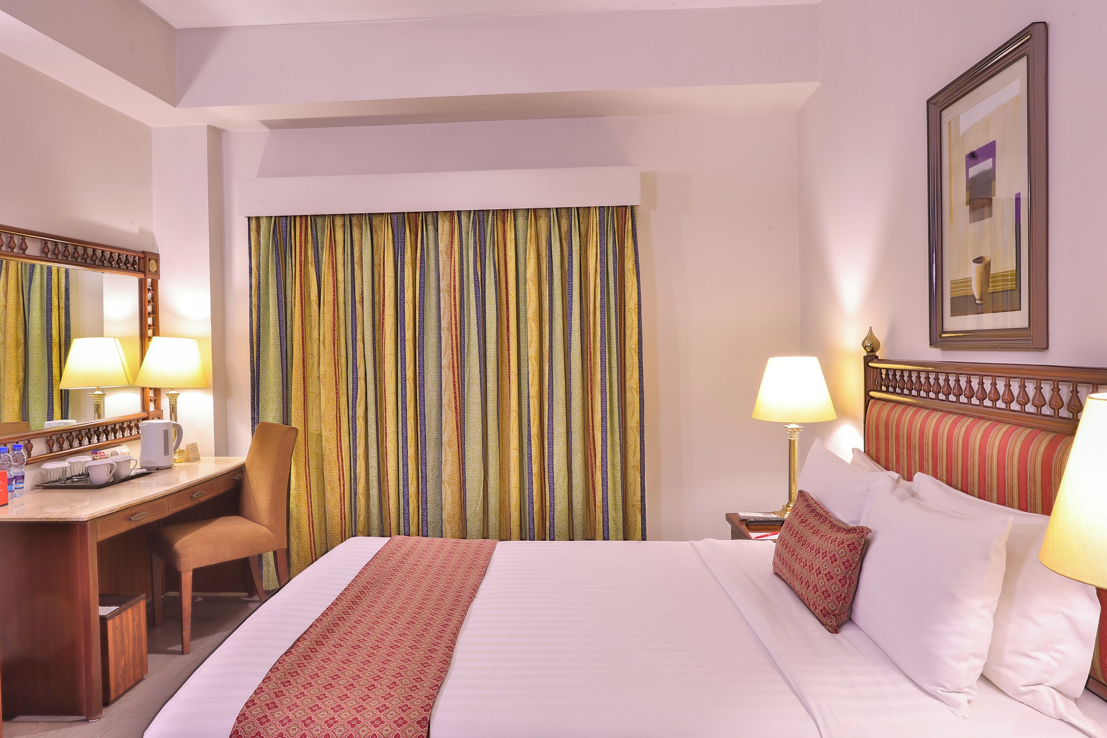 Habitación de hotel con cama, escritorio y silla, lámpara de escritorio, mesita de noche con lámpara y un ventanal con cortinas.