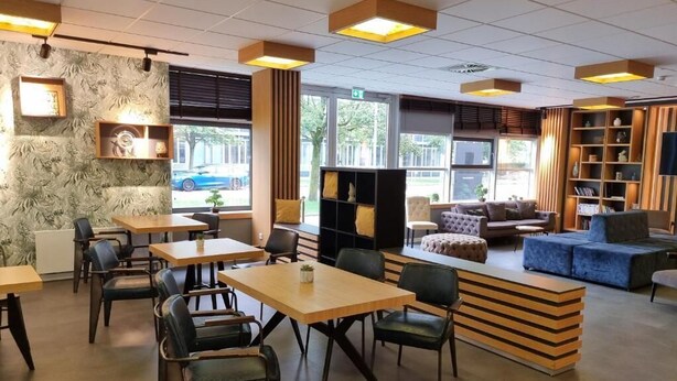 Una cafeteria moderna amb taules i cadires de fusta, una zona amb sofà i una prestatgeria de llibres.