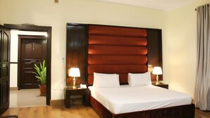 A hotel room with a large bed, two bedside tables with lamps, a wooden headboard, a potted plant, and a door with a mirror. - Sunset Destination (Islamabad)