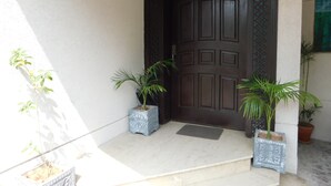 A dark wooden door with a decorative panel, flanked by two potted plants on a tiled porch. - Sunset Destination (Islamabad)