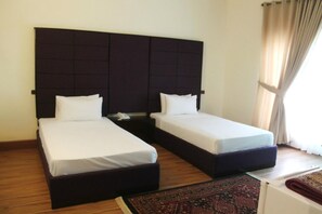 A hotel room with two single beds, a dark headboard, a small bedside table with a phone, and a window with curtains. - Sunset Destination (Islamabad)