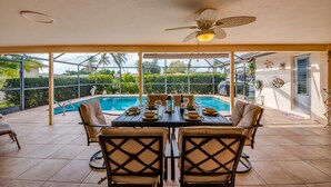 Un patio iluminado por el sol con piscina, mesa de comedor y sillas, con vistas a un jardín ajardinado.