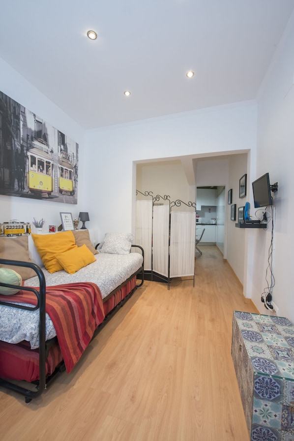 A compact bedroom with a bed, a small desk, and a kitchenette in the background.