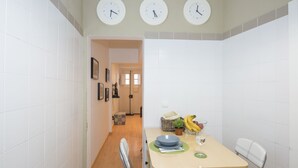 Una cocina pequeña y luminosa con azulejos blancos, tres relojes de pared, una mesa de comedor con sillas y un vano que da a otra habitación.