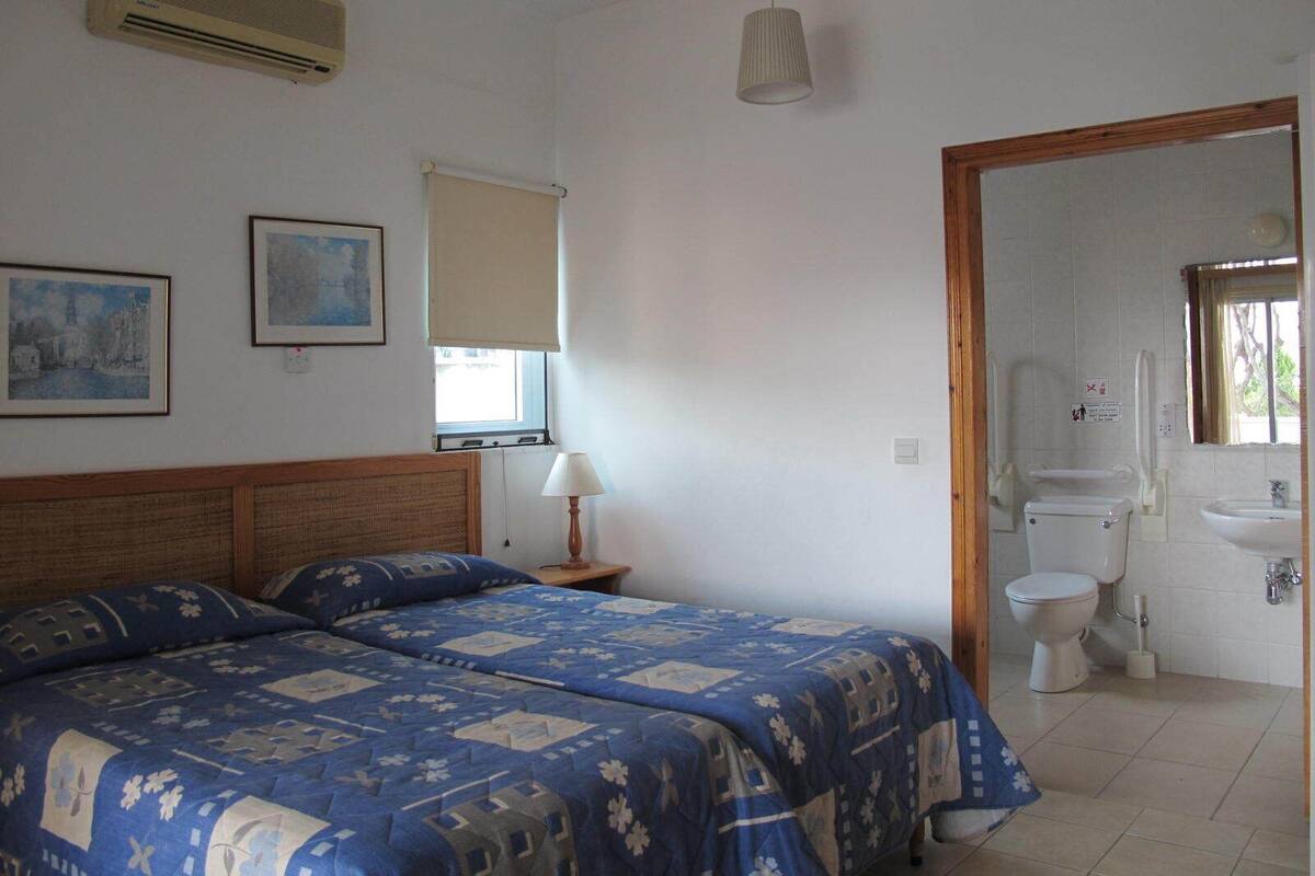 A bedroom with two beds, a window with a blind, a bedside lamp, and a bathroom visible through an open door.