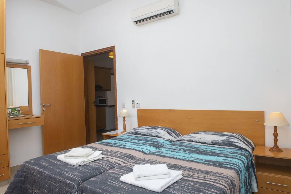 A hotel room with a bed, a nightstand, a lamp, a mirror, a wardrobe, and an air conditioning unit.