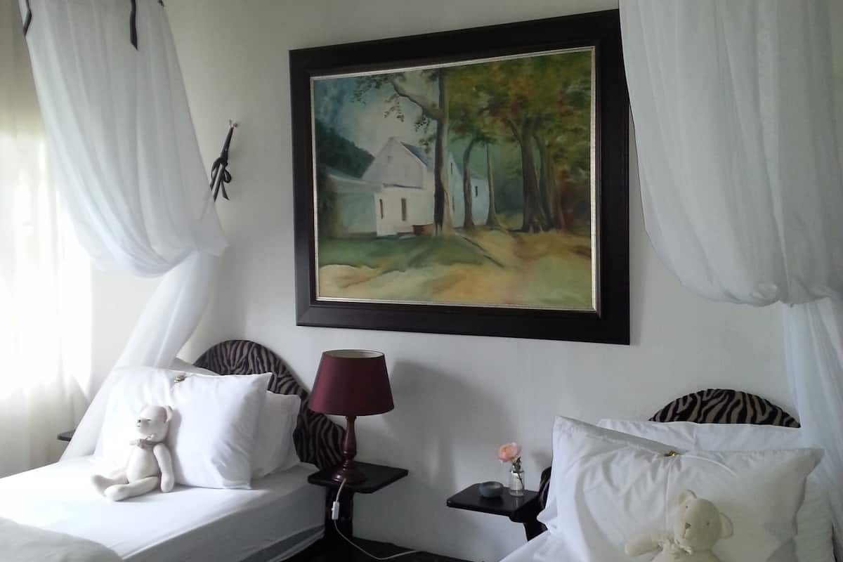 Une chambre d’hôtel avec deux lits, chacun agrémenté d’un oreiller blanc et d’un jouet en peluche. Un tableau est accroché au mur au-dessus des lits, il y a une petite table avec une lampe, et un vase avec une fleur sur la table de chevet.
