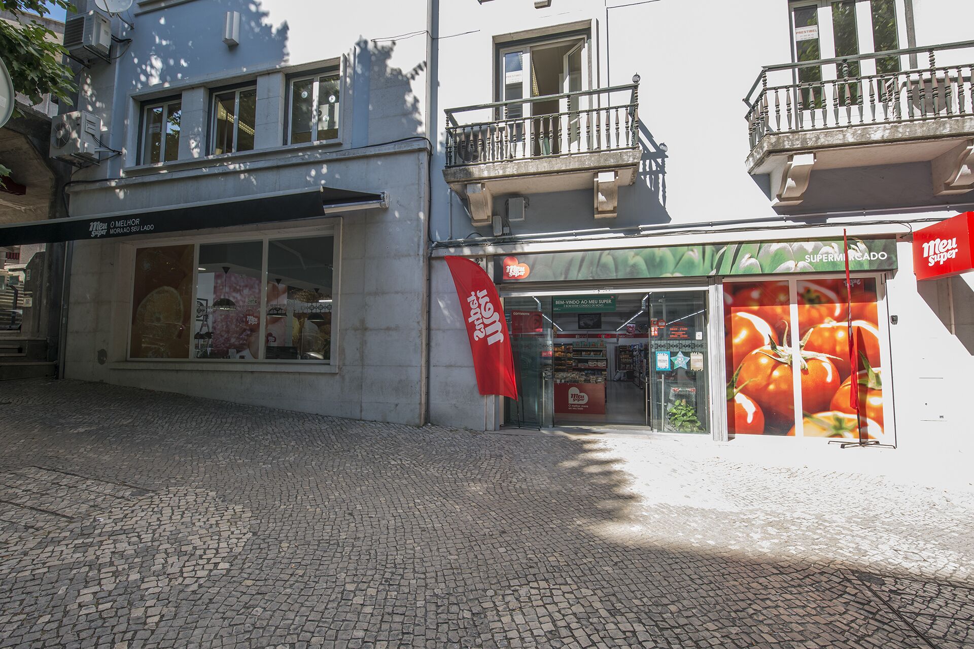 Une devanture de magasin avec une grande vitrine où sont exposées des tomates, une banderole rouge et une enseigne indiquant « Meu Super ».