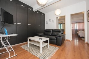 Una sala de estar moderna con un sofá de cuero negro, una mesa de centro blanca y un televisor montado en la pared. Hay un escritorio con una computadora y una lámpara. El piso es de madera y hay un ventanal grande con cortinas.