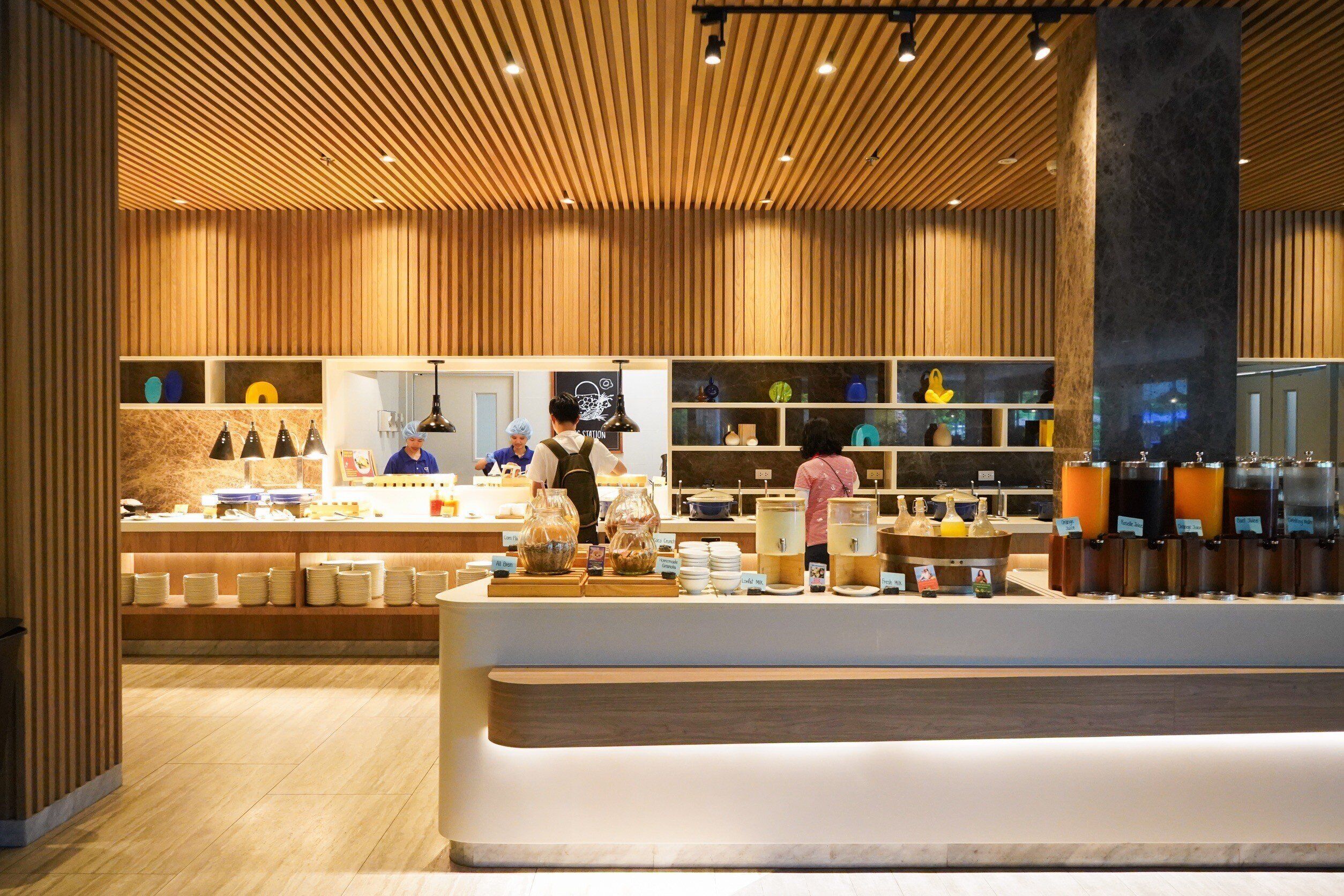 A modern cafe with wooden paneling, a counter with various food items, and a barista preparing drinks.