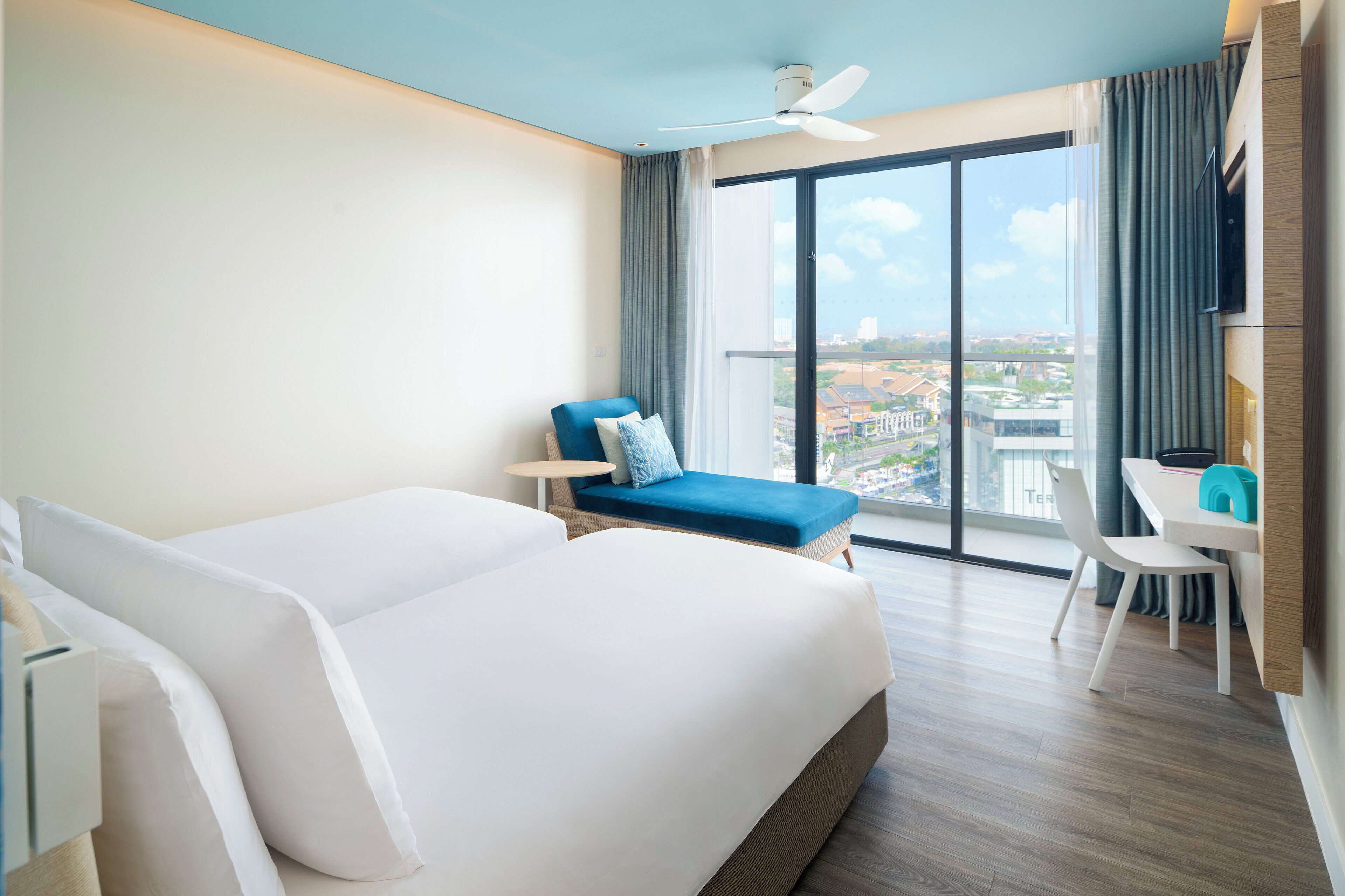 Een hotelkamer met een groot bed, een blauwe fauteuil, een bureau en uitzicht op de stad door grote ramen.