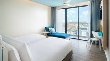 Een hotelkamer met een groot bed, een blauwe fauteuil, een bureau en uitzicht op de stad door grote ramen.
