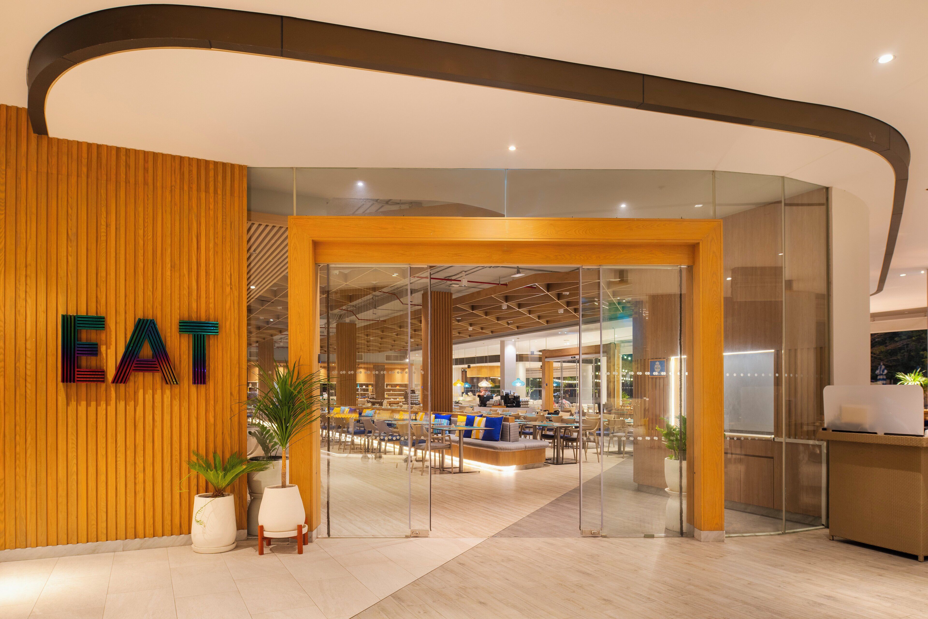 A modern eatery with a wooden wall and a glass entrance. Inside, there are tables and chairs arranged for dining.