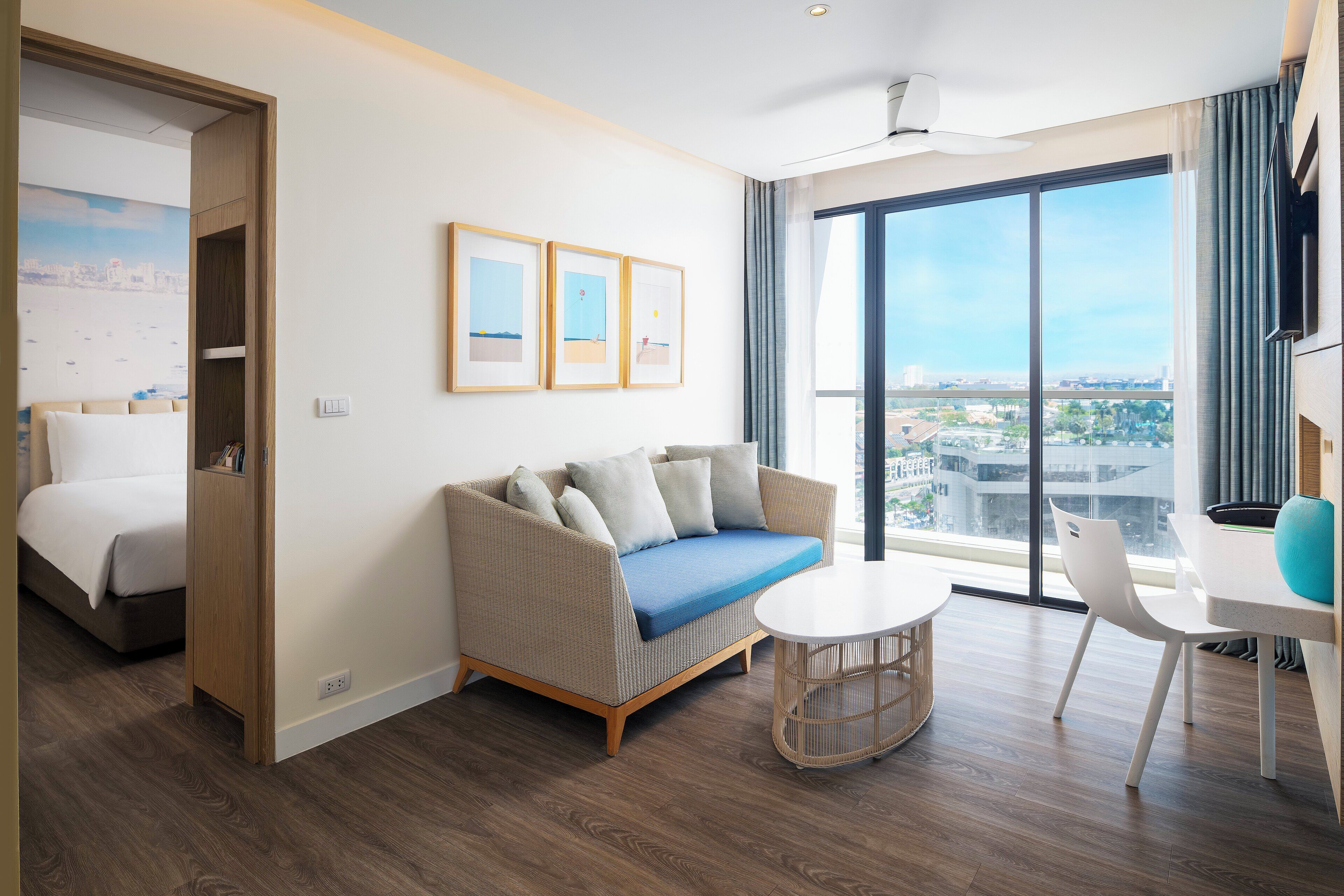 A modern hotel room with a bed, a sofa, a dining table, and a large window offering a city view.