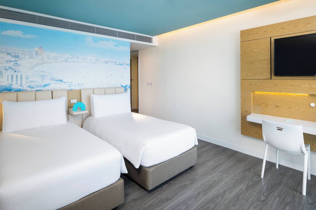 Habitación de hotel con dos camas, un escritorio, una silla, un televisor montado en la pared y un mural con vista a la ciudad en el techo.