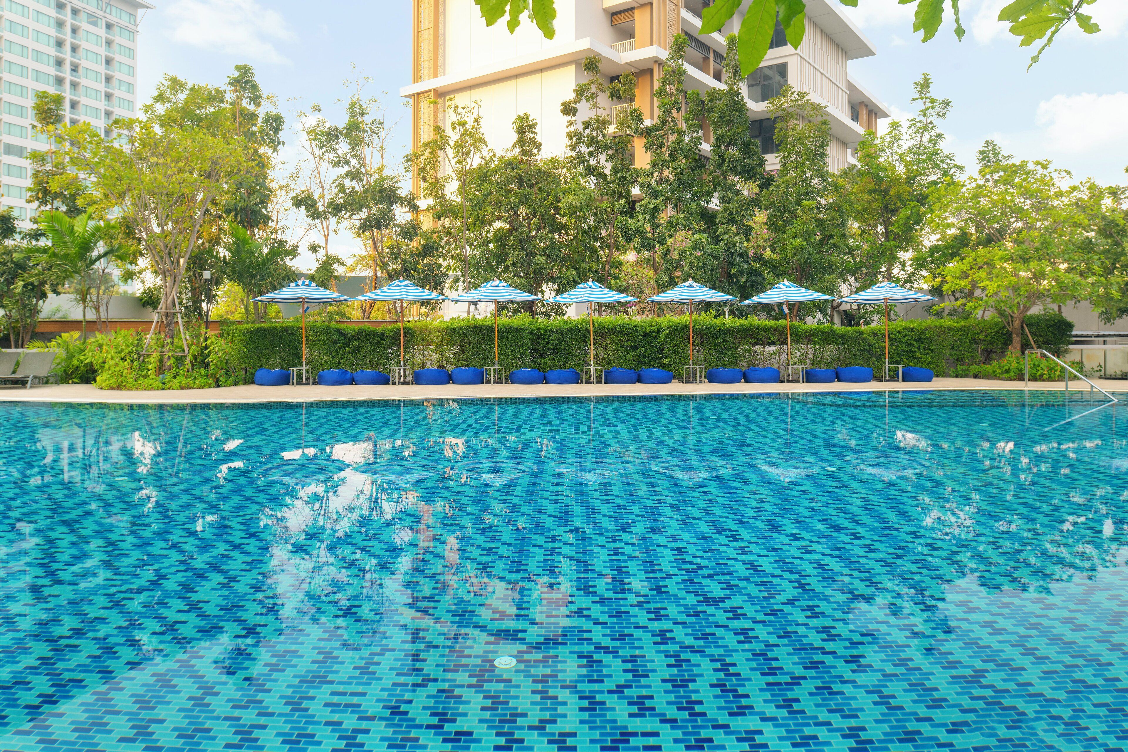 Una piscina con piso de azulejos azules, rodeada de sillas de descanso y sombrillas azules, con árboles y un edificio al fondo.