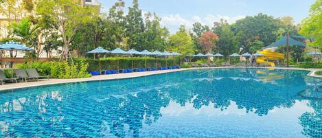 A swimming pool with tiled blue and green patterns, surrounded by lounge chairs and umbrellas, with trees and a building in the background.