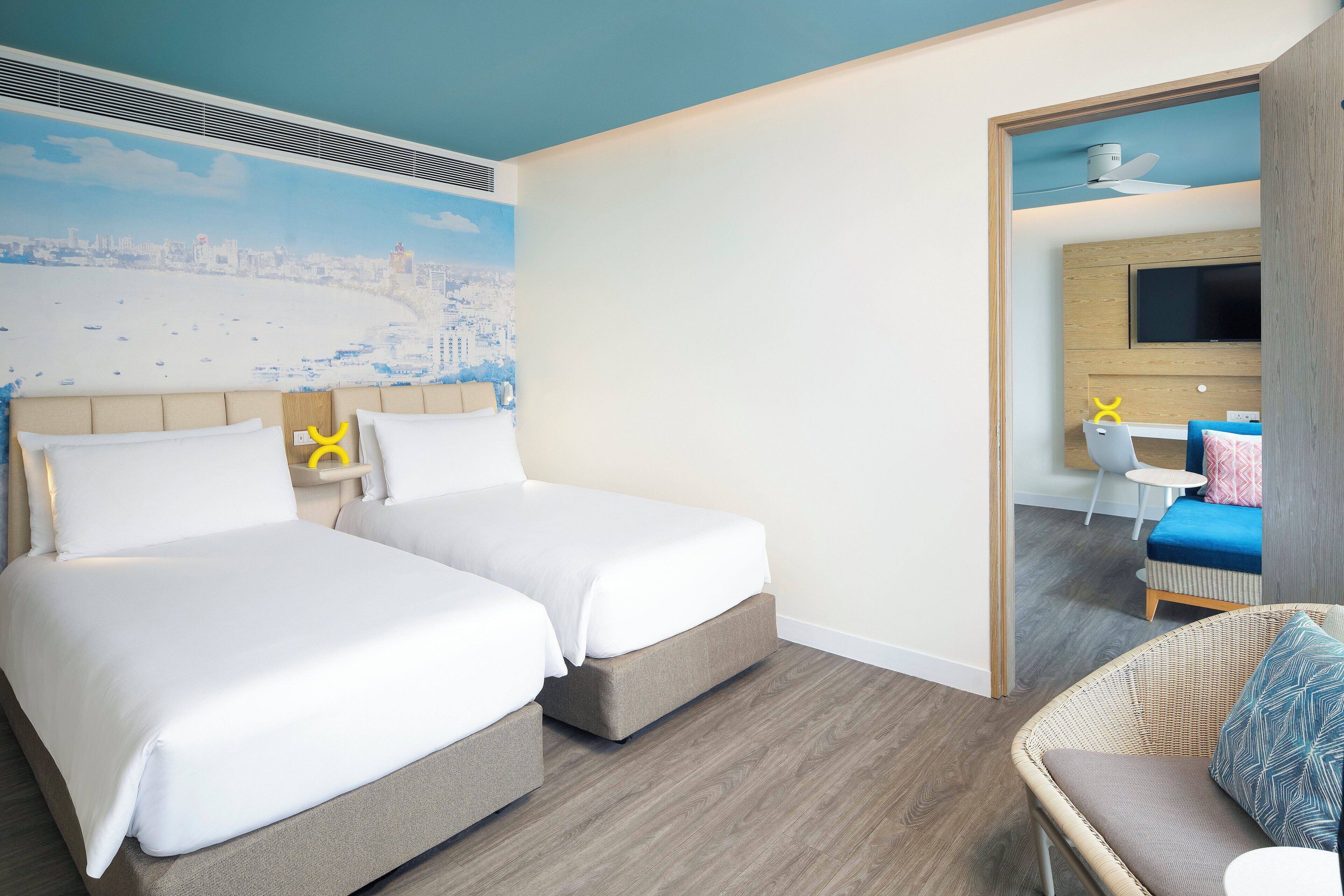 Habitación de hotel con dos camas, un mural de la ciudad, un televisor y una silla de mimbre.