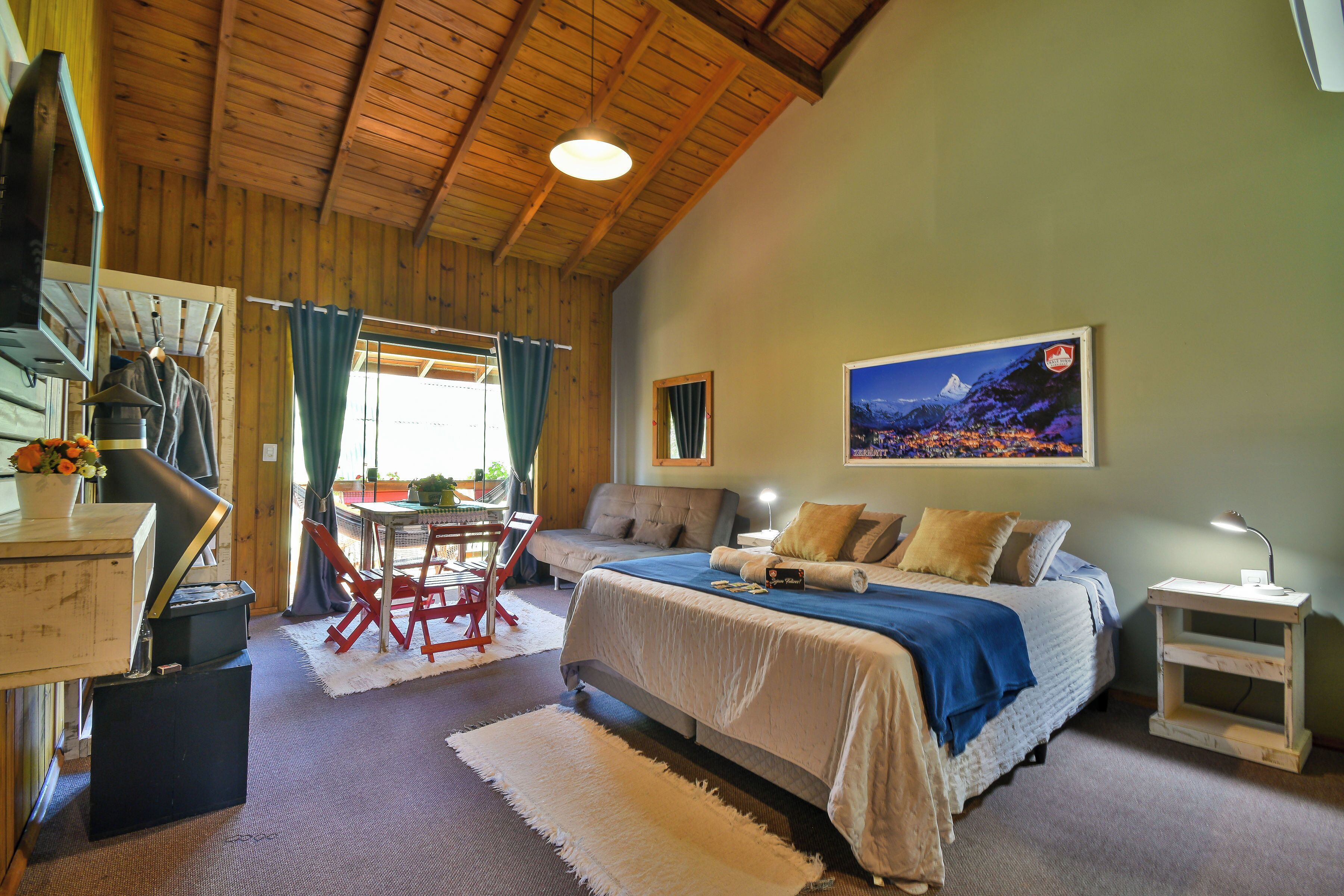 Une pièce chaleureuse avec un plafond en bois, un lit recouvert d’une couverture bleue, un bureau avec une chaise et un tableau accroché au mur.