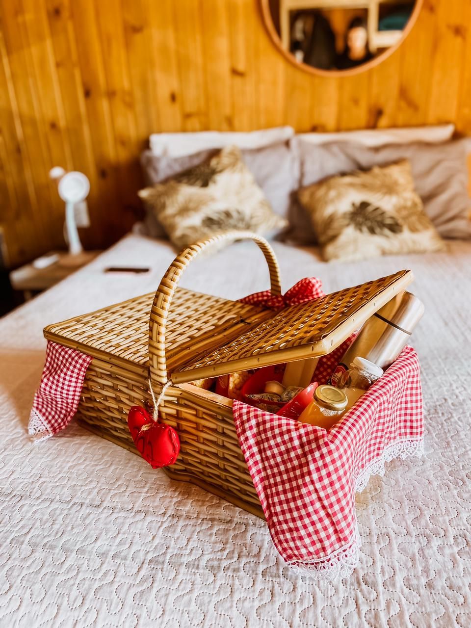 A wicker basket with a red and white checkered cloth, containing a bottle, a jar, and a red ornament, placed on a bed with a white textured cover.
