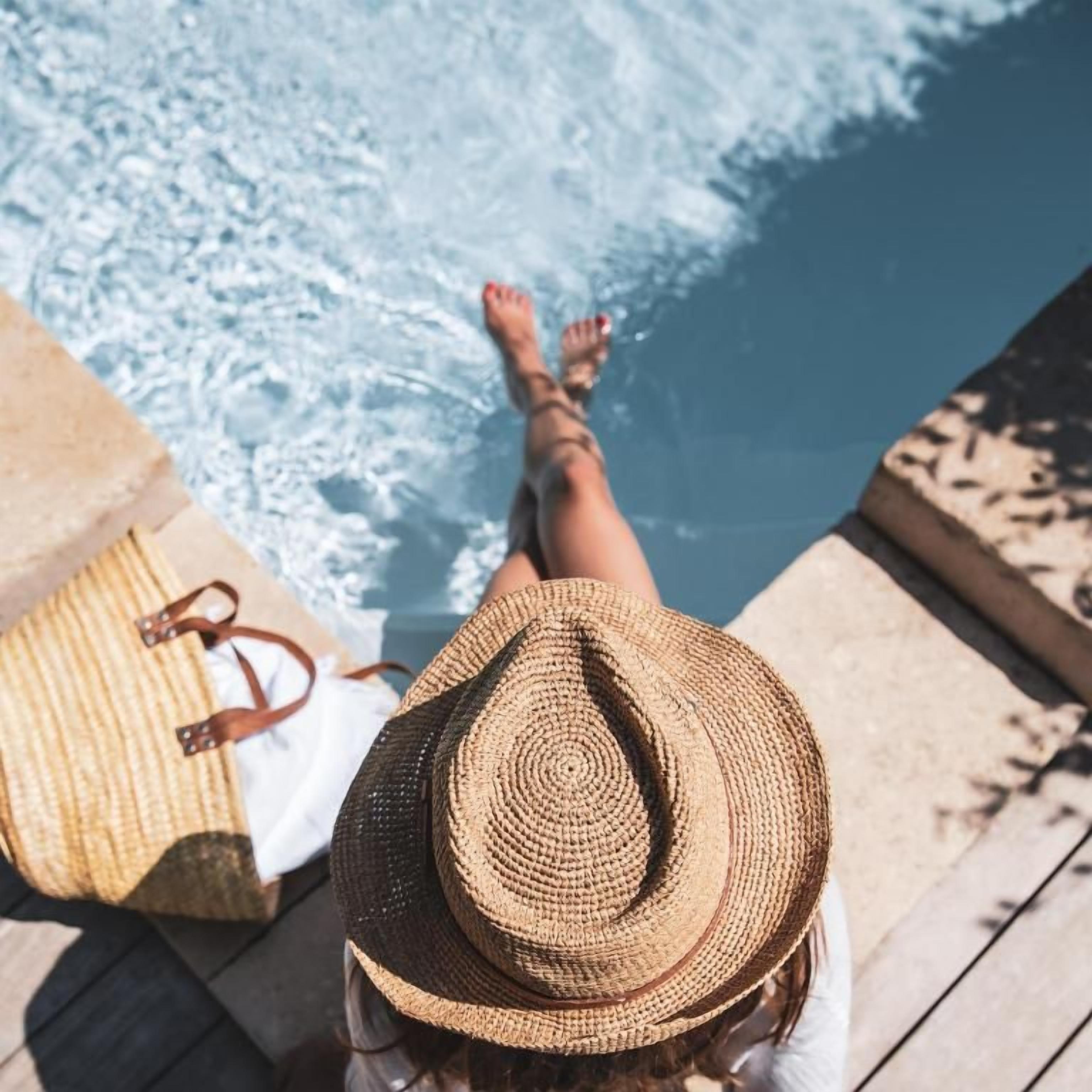 A person sitting by a pool with a straw hat, a beach bag, and a wooden deck.