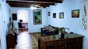 A cozy living room with a wooden coffee table, a sofa, and a desk with a computer. There are several framed pictures on the walls and a ceiling fan above. - Deck Hostel Congonhas (São Paulo)