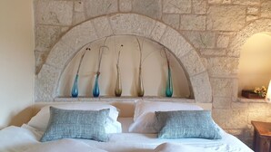 Un lit avec du linge de lit blanc et deux oreillers gris, un mur de pierre avec une niche en arc de cercle, et cinq vases de différentes couleurs.