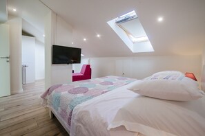 A bedroom with a skylight, a bed with white bedding, a pink chair, and a flat-screen TV mounted on the wall.