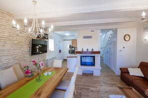 A modern living room with a wooden dining table, a fireplace, a brown leather sofa, and a chandelier.