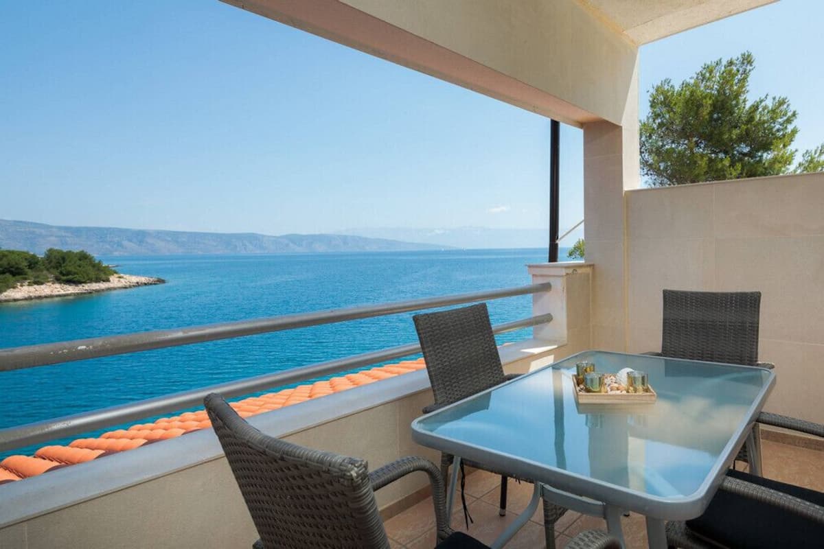 A balcony with a glass table and wicker chairs, overlooking a body of water and a distant coastline.