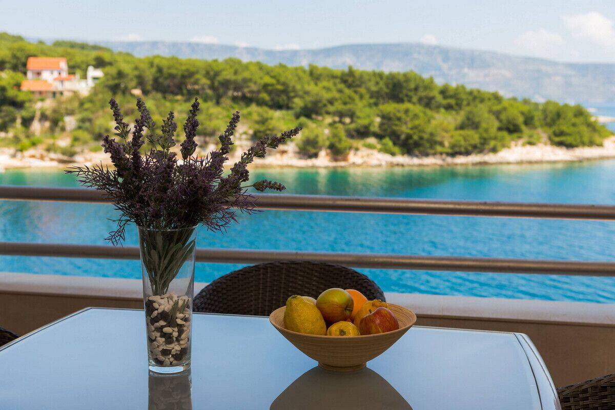 A table with a vase of flowers and a bowl of fruit by a poolside with a view of a beach and mountains.
