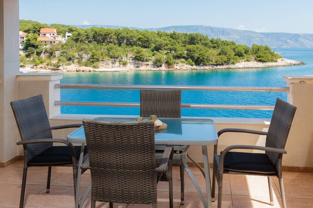 A balcony with a view of a turquoise sea and a green hillside. There are wicker chairs and a glass table.