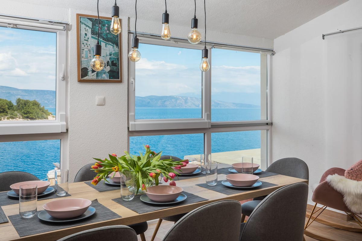 A dining area with a view of the sea, a table set with pink bowls and plates, and a vase with flowers.