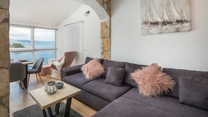A modern living room with a stone archway, a large window with a view of the sea, a gray sofa with fluffy pillows, and a painting of sailboats on the wall. - Apartments Habek (Jelsa)