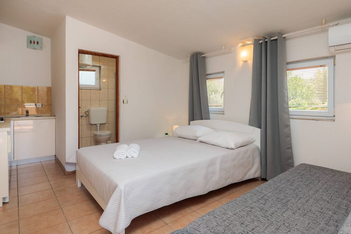 A compact hotel room with a bed, a small kitchenette, a bathroom, and a window with curtains.
