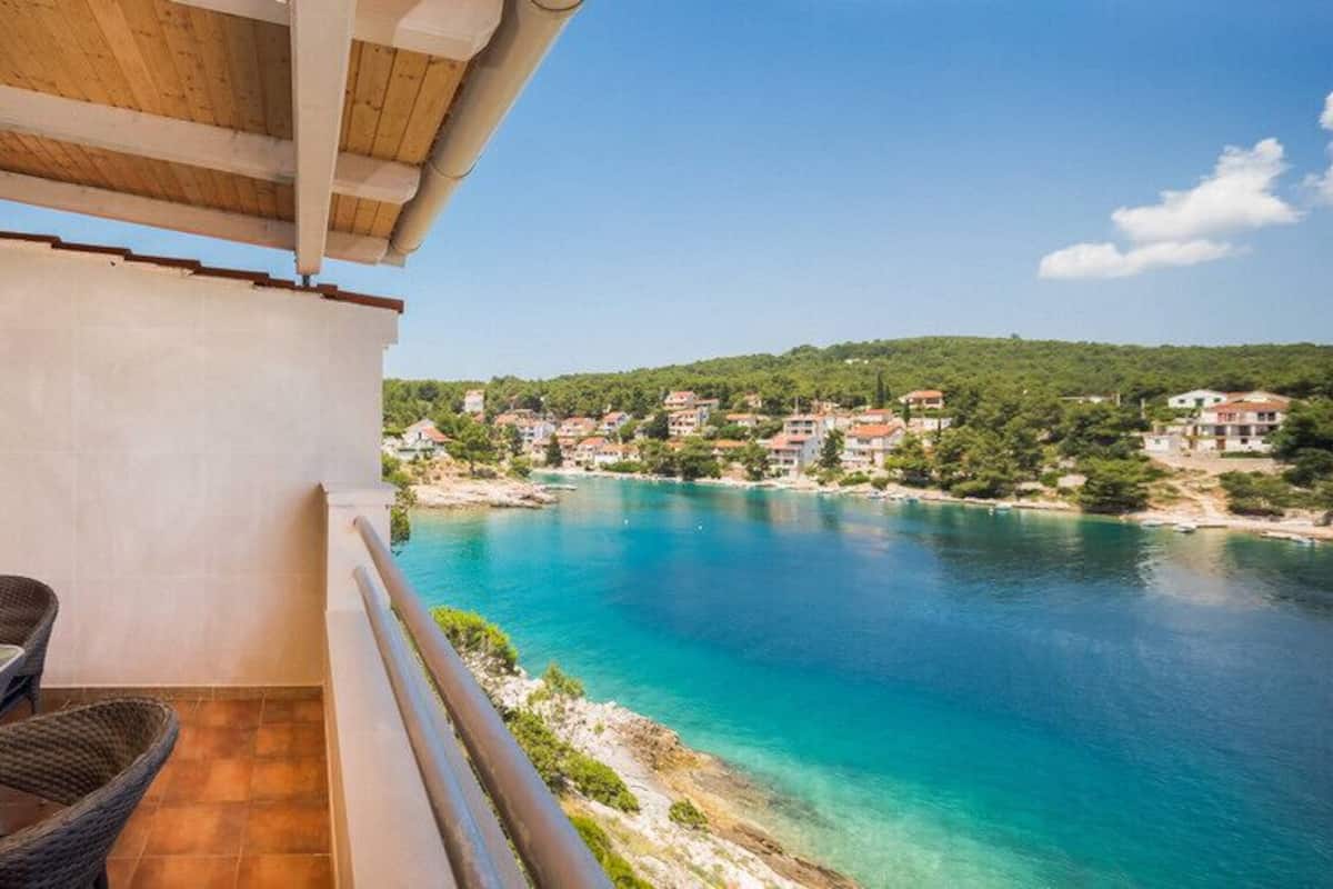 A balcony with a table and chairs overlooking a turquoise sea and a coastal village.