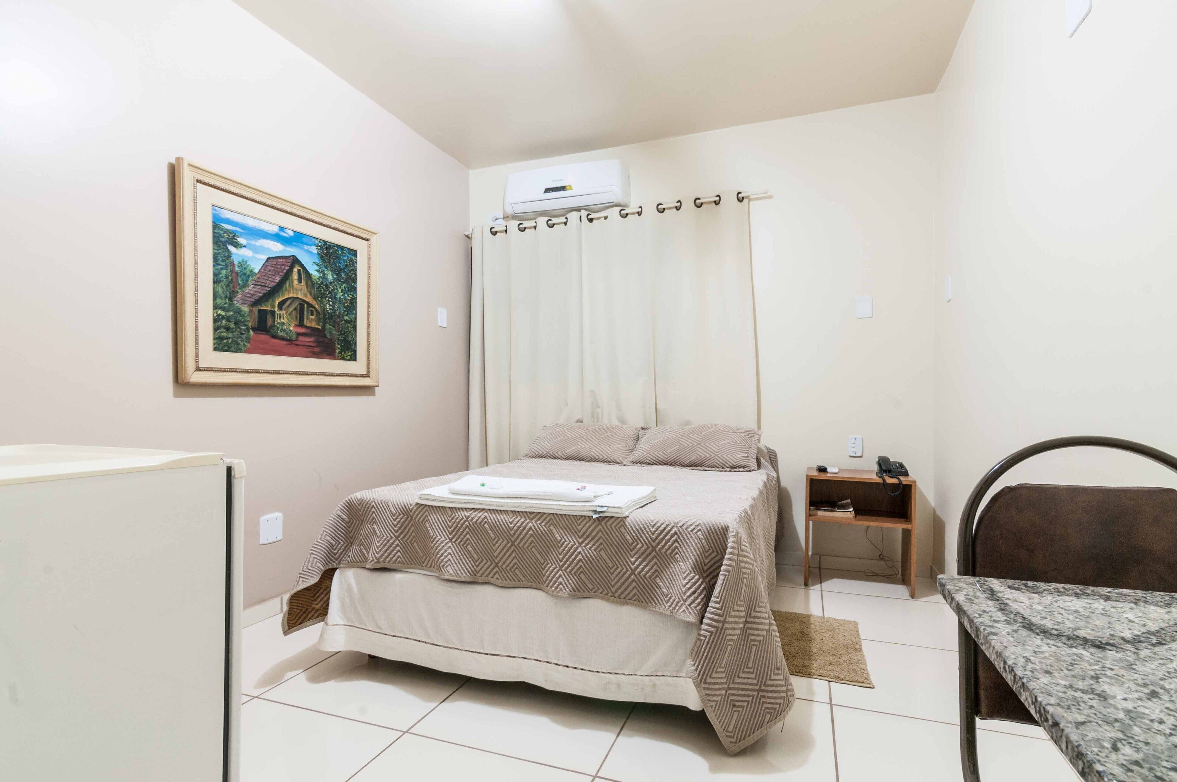 Quarto com cama, mesa de cabeceira, escrivaninha, cadeira, quadro na parede e ar condicionado.